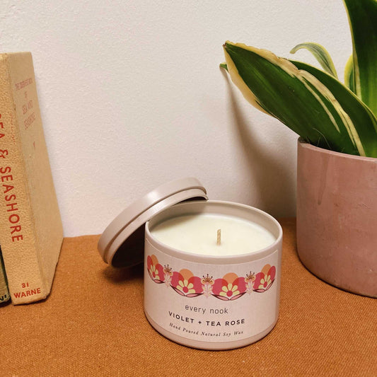 Candle in a decorative tin with floral design on a table next to a book and plant